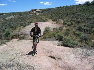 A mountain biker navigating a rocky trail surrounded by sparse vegetation and blue sky with white clouds. Hartman Rocks mountain bike trail.