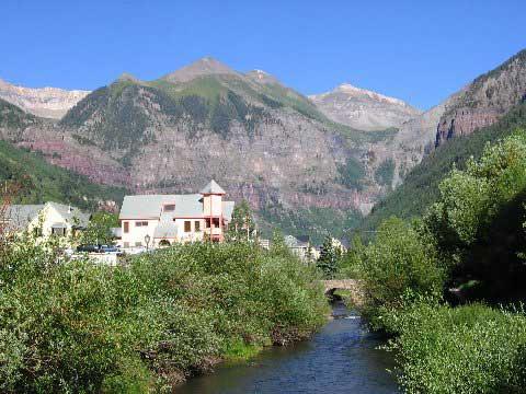 A scenic view of a mountain landscape featuring a clear blue sky, a gently flowing stream, and a few buildings nestled among lush greenery at the base of the mountains. River Trail mountain bike trail.