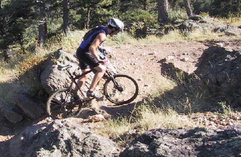 A cyclist riding a mountain bike on a rocky trail surrounded by trees and grass. White Ranch mountain bike trail.