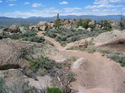 A winding dirt trail traversing a rocky landscape, surrounded by sparse vegetation and shrubs, with distant mountains against a blue sky dotted with clouds. Hartman Rocks mountain bike trail.