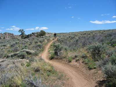 A mountain bike trail winding through a grassy landscape under a clear blue sky. A cyclist is visible in the distance on the sandy path, surrounded by sparse vegetation and rocky terrain. Hartman Rocks mountain bike trail.