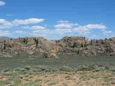 A scenic view of rocky outcrops surrounded by a grassy plain under a bright blue sky with scattered clouds. The landscape features sparse vegetation and hints of evergreen trees among the rugged terrain. Hartman Rocks mountain bike trail.