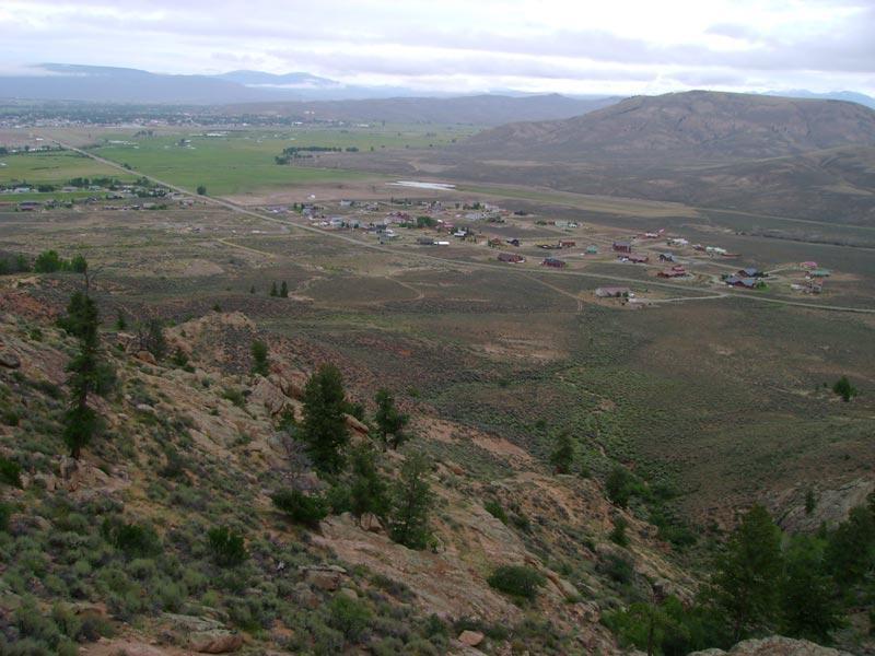 A panoramic view of a small town nestled in a valley, surrounded by rolling hills and mountains. The foreground features rocky terrain and sparse vegetation, while the background showcases lush green fields and scattered residential buildings. Overcast skies hover above, adding a serene atmosphere to the landscape. Hartman Rocks mountain bike trail.
