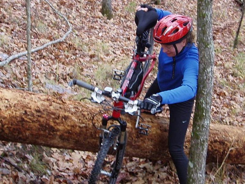 A person in a blue long-sleeve shirt and a red helmet is attempting to lift a mountain bike over a fallen log in a wooded area covered with autumn leaves. The scene captures the challenge of navigating rough terrain while biking. Hickory Knob State Park mountain bike trail.