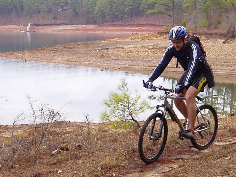 A man in a cycling outfit rides a mountain bike along a dirt path near a body of water, with a forested area and sandy shoreline in the background. Hickory Knob State Park mountain bike trail.