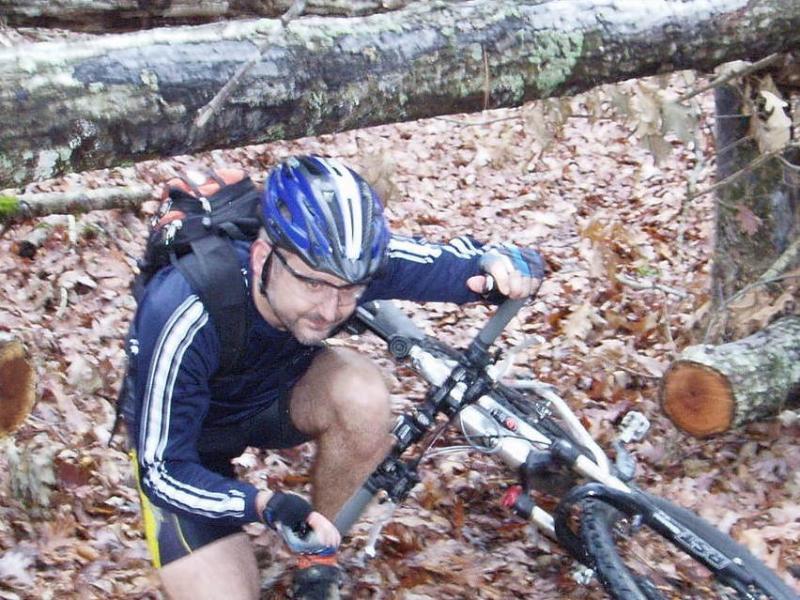 A mountain biker crouching next to a bike in a forested area, navigating around fallen logs and surrounded by fallen leaves. The cyclist is wearing a blue helmet and a dark athletic outfit, focused on maneuvering the bike. Hickory Knob State Park mountain bike trail.