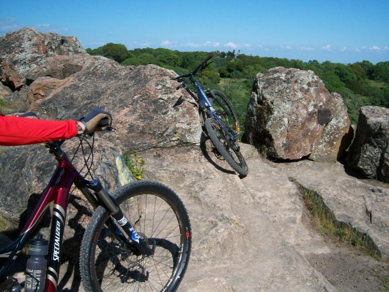A close-up view of a mountain bike resting on rocks with a scenic landscape in the background. One bike is positioned against large boulders, while another bike is held by a person wearing a red long-sleeve shirt. The sky is clear with a few clouds, and greenery is visible in the distance. Rockville Park mountain bike trail.