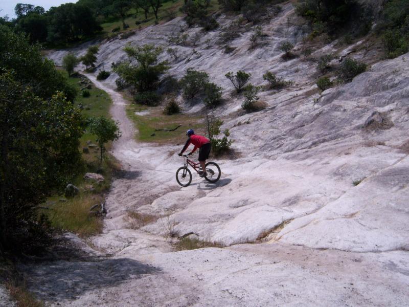 A mountain biker navigates a rocky, sloped terrain surrounded by sparse vegetation and trees. A dirt path is visible in the background, leading further into the landscape. Rockville Park mountain bike trail.