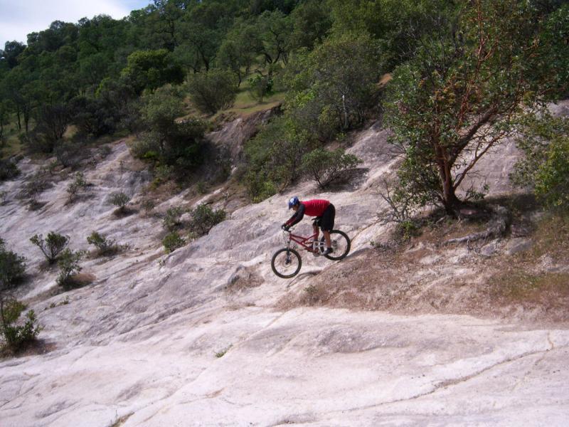 Mountain biker navigating a rocky terrain, with a focus on the steep slope and surrounding vegetation. The cyclist wears a helmet and red clothing and appears to be skillfully maneuvering the bike down the rocky surface. Rockville Park mountain bike trail.