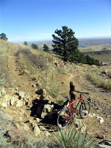 A red mountain bike is positioned next to a rustic wooden post on a rocky trail overlooking a vast valley and distant hills. The scene features sparse vegetation, including a few trees, under a clear blue sky. White Ranch mountain bike trail.