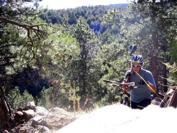 A person wearing a helmet and sunglasses is pushing a mountain bike up a rocky trail surrounded by lush green pine trees. Bright sunlight filters through the trees, illuminating the scenic landscape in the background. White Ranch mountain bike trail.