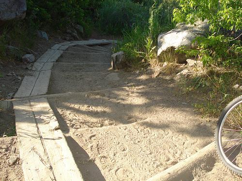 A natural pathway made of wooden planks leading down a sloped, sandy area, surrounded by greenery and large rocks. A bicycle is partially visible on the right side of the image. The scene is illuminated by warm sunlight, casting soft shadows on the ground. White Ranch mountain bike trail.