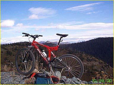 A red mountain bike is positioned on a rocky outcrop with a panoramic view of rolling hills and a blue sky in the background. Tree-covered mountains stretch into the distance, and the scene captures a sense of adventure and outdoor exploration. Whitefish Mountain Resort mountain bike trail.