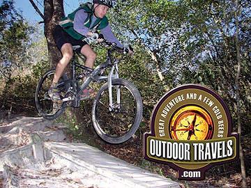 A mountain biker mid-jump on a wooded trail, showcasing an outdoor adventure. The image includes a logo for "Outdoor Travels" with the tagline "Great adventure and a few cold beers." Alafia River State Park mountain bike trail.