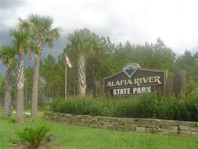 Signage at Alafia River State Park, featuring palm trees and lush greenery, under a cloudy sky. The park's name is prominently displayed, along with a park emblem and a flag. Alafia River State Park mountain bike trail.
