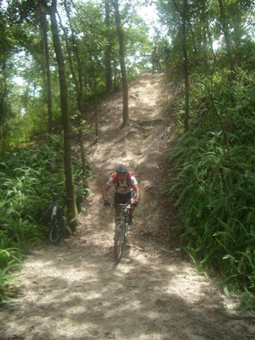 Mountain biker pushing up a sandy hill surrounded by trees and lush greenery. Another bike is resting on the trail in the background. Alafia River State Park mountain bike trail.