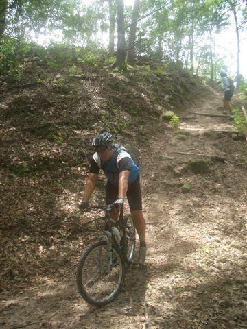 A person in athletic clothing pushes a mountain bike up a dirt path in a wooded area, surrounded by trees and foliage. The path is narrow and steep, with sunlight filtering through the leaves. Alafia River State Park mountain bike trail.