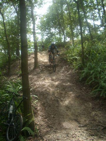 A mountain biker descending a dirt trail surrounded by trees and vegetation, with another bicycle placed on the left side of the image. Alafia River State Park mountain bike trail.