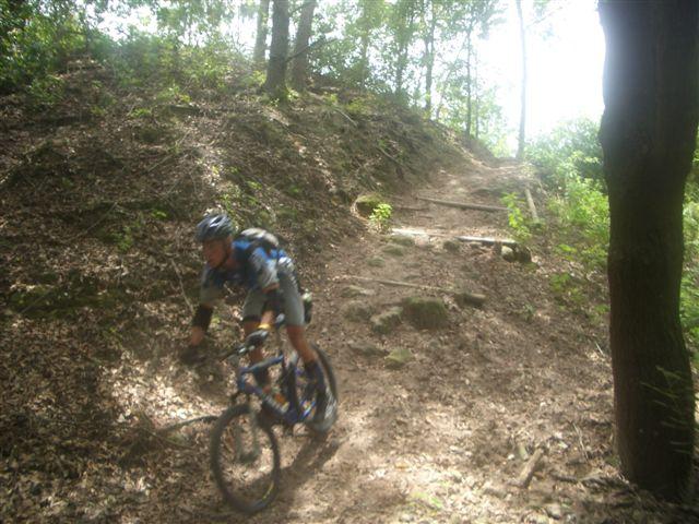 Mountain biker navigating a rocky, wooded trail, with a slope leading upwards in the background. The cyclist is wearing a helmet and riding a blue bike, surrounded by greenery and sunlight filtering through the trees. Alafia River State Park mountain bike trail.