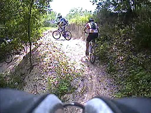 Three mountain bikers navigating a dirt trail surrounded by greenery. The trail includes a hill that the riders are climbing, with varying terrain visible. The scene captures an outdoor cycling activity. Alafia River State Park mountain bike trail.