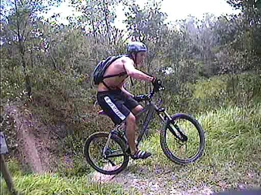 A person riding a mountain bike on a trail, wearing a helmet and a backpack, as they prepare to jump off a small incline surrounded by greenery. Alafia River State Park mountain bike trail.