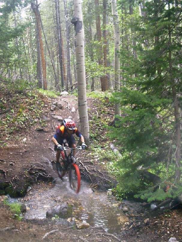 A mountain biker rides over a rocky trail, splashing through a small stream while surrounded by lush green trees in a forested area. The rider wears a helmet and is in an action pose, showcasing the thrill of off-road biking. Monarch Crest Trail mountain bike trail.