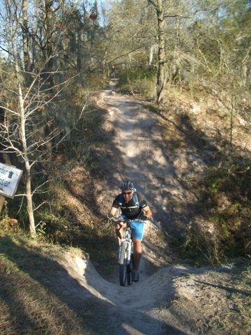 A cyclist navigating a dirt bike trail, pushing their mountain bike uphill. The path is lined with trees and foliage, under a clear blue sky. The terrain appears rugged and natural, indicative of a well-used biking trail. Alafia River State Park mountain bike trail.