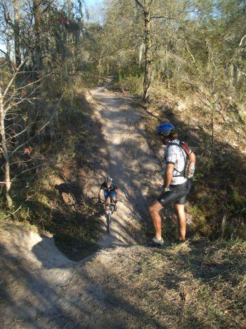 A mountain biker navigating a dirt trail while another person stands on the side, observing. The scene is set in a wooded area with trees in the background and a clear sky. The path is uneven and winding, suggesting an outdoor adventure. Alafia River State Park mountain bike trail.