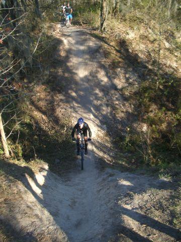 A mountain biker navigating a dirt trail surrounded by trees. The trail has a steep slope and a winding path, with shadows cast from the trees above. Another cyclist can be seen in the background, riding on a separate part of the trail. Alafia River State Park mountain bike trail.