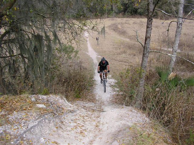 A mountain biker riding down a dirt trail surrounded by trees and sparse grassland. The trail winds through the landscape, and a glimpse of another person can be seen in the distance. Alafia River State Park mountain bike trail.