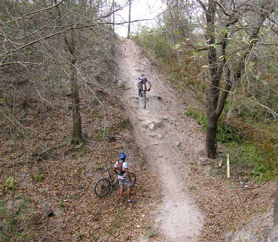 Two mountain bikers navigating a rocky trail through a wooded area with dry leaves on the ground. One biker is ascending the trail, while the other is standing with their bike, observing the terrain. The surroundings are sparse with trees, and the trail is steep and uneven. Alafia River State Park mountain bike trail.