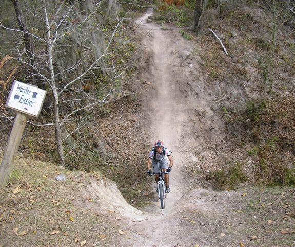 A mountain biker navigating a dirt trail that slopes downward, with a signpost to the left indicating a choice between "Harder" and "Easier" paths. The landscape features sparse trees and autumn foliage. Alafia River State Park mountain bike trail.