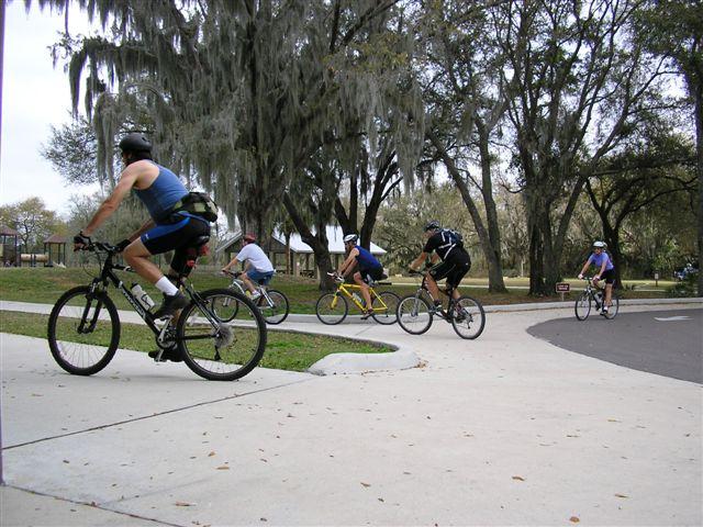 A group of six cyclists riding on a paved path through a park, surrounded by trees and natural scenery. The riders are dressed in athletic gear and are seen navigating a curve in the path. Alafia River State Park mountain bike trail.