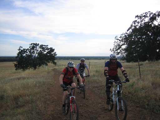 Three mountain bikers riding along a dirt path in a grassy landscape, with trees and a distant view of rolling hills under a cloudy sky. Upper Bidwell Park mountain bike trail.