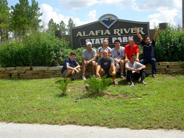 A group of eight people posing in front of the Alafia River State Park sign, surrounded by greenery and a sunny sky. Some individuals are sitting and others are standing, with expressions of joy and camaraderie. Alafia River State Park mountain bike trail.
