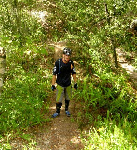 A cyclist standing on a dirt trail surrounded by lush greenery and trees, wearing a helmet, gloves, and biking gear. Alafia River State Park mountain bike trail.