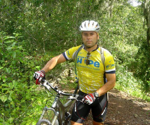 A male mountain biker wearing a yellow and blue cycling jersey stands beside his bike on a dirt trail surrounded by greenery. He has a helmet on and is looking directly at the camera, showcasing a confident expression. The background features lush trees and foliage typical of a forested area. Alafia River State Park mountain bike trail.
