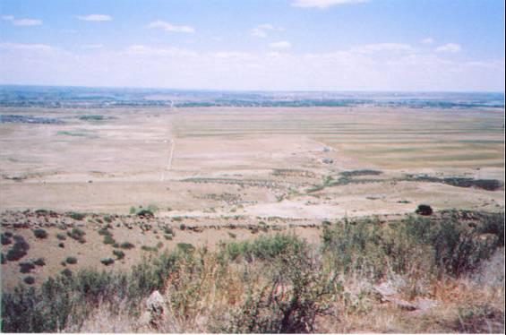 A wide view of a dry, expansive landscape featuring arid fields and patches of greenery in the foreground. The scene is set under a bright blue sky with scattered clouds, stretching out to the horizon where distant hills can be seen. A faint road is visible winding through the landscape. Coyote Ridge mountain bike trail.