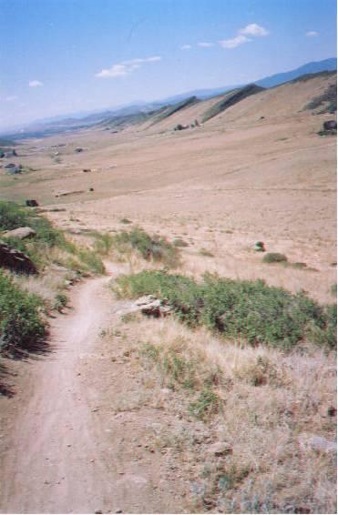 A winding dirt trail leading down a grassy hillside, with expansive dry fields and distant mountains under a blue sky scattered with clouds. Coyote Ridge mountain bike trail.