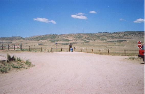 A dirt road leading towards an open landscape with rolling hills under a bright blue sky. A wooden fence lines the road, and a stop sign is visible on the right side near a red vehicle. A small information booth can be seen in the distance, surrounded by sparse vegetation. Coyote Ridge mountain bike trail.