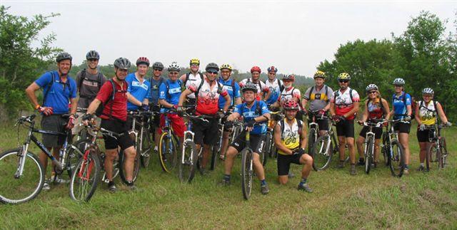 A group of 20 cyclists wearing helmets and colorful biking attire pose together with their mountain bikes on a grassy area, surrounded by trees. They are smiling and appear to be enjoying a biking event or outing. Alafia River State Park mountain bike trail.