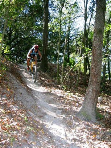 A mountain biker navigating a winding dirt trail through a wooded area, surrounded by trees and fallen leaves, with sunlight filtering through the foliage. Alafia River State Park mountain bike trail.