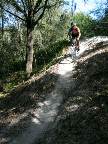 A mountain biker descending a sandy trail in a wooded area, surrounded by greenery and trees. Alafia River State Park mountain bike trail.
