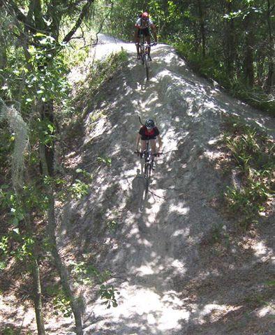 Two mountain bikers riding on a dirt trail through a wooded area, one descending a sandy incline while the other is further up the trail. Sunlight filters through the trees, casting shadows on the ground. Alafia River State Park mountain bike trail.