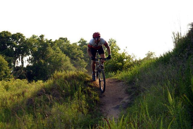 Mountain biker navigating a dirt trail on a sunny day, surrounded by greenery and hills. Alafia River State Park mountain bike trail.