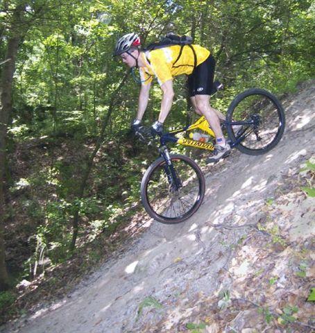 A mountain biker in a yellow shirt and helmet riding down a steep, dirt slope surrounded by trees in a forested area. Alafia River State Park mountain bike trail.