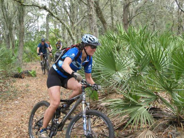 A woman rides a mountain bike on a wooded trail, surrounded by greenery and palm plants. She wears a blue cycling jersey and a helmet. In the background, two other cyclists can be seen following the trail. Alafia River State Park mountain bike trail.