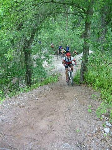 A mountain biker navigating a downhill trail surrounded by lush green trees, with other cyclists visible in the background. The terrain is sandy and is situated in a wooded area, showcasing a recreational outdoor activity. Alafia River State Park mountain bike trail.