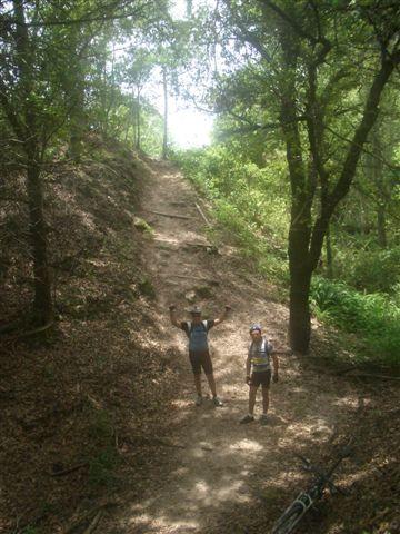 Two hikers standing on a dirt trail in a wooded area, surrounded by trees and lush greenery. One hiker has arms raised in celebration, while the other smiles at the camera. Sunlight filters through the trees, creating a bright and inviting atmosphere. Alafia River State Park mountain bike trail.