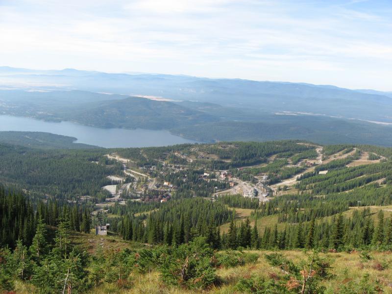 A panoramic view of a mountainous landscape featuring thick green pine forests, a winding road, and a serene lake in the background. The sky is clear with a few clouds, enhancing the natural beauty of the scene. The foreground showcases vibrant vegetation, while the middle ground reveals a developed area with buildings and ski slopes, indicative of a recreational destination. Whitefish Mountain Resort mountain bike trail.